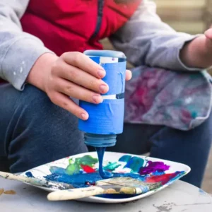 A kid squeezing blue paint onto a white plate with brushes and other colors.